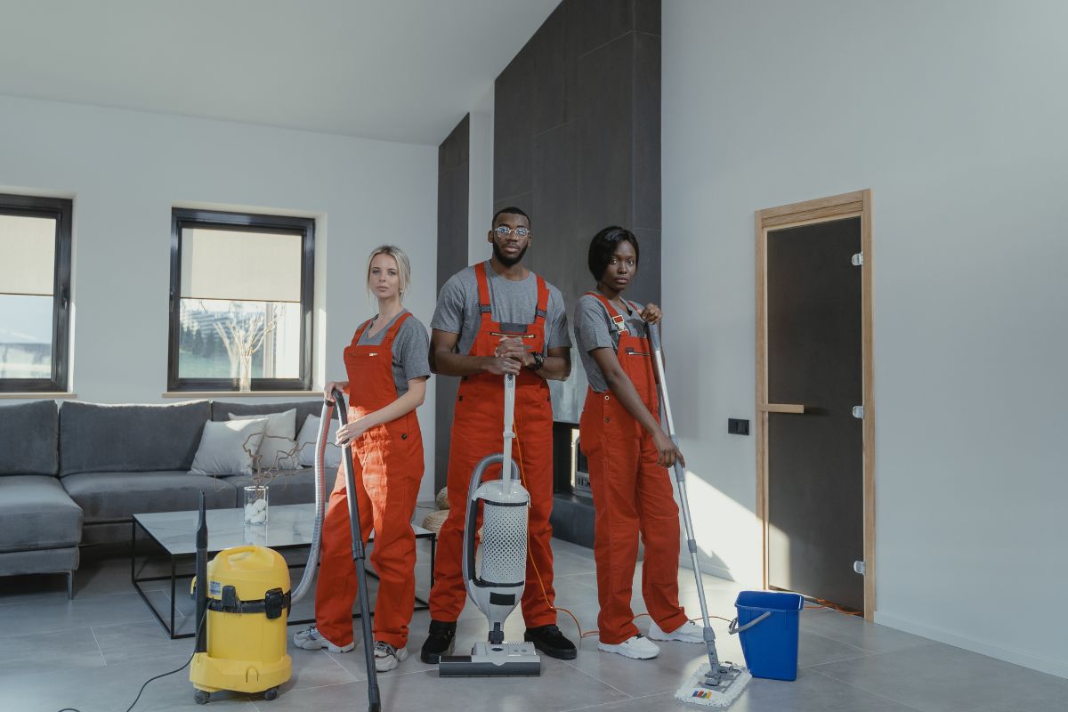 Three individuals in overalls stand in a room surrounded by various vacuum cleaners, showcasing a cleaning equipment display.