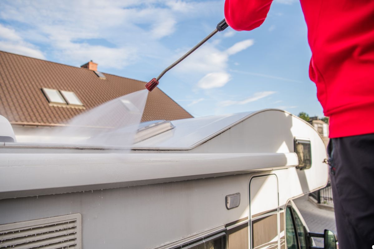 A man scrubs the roof of a recreational vehicle, standing on a ladder with cleaning supplies in hand. 
