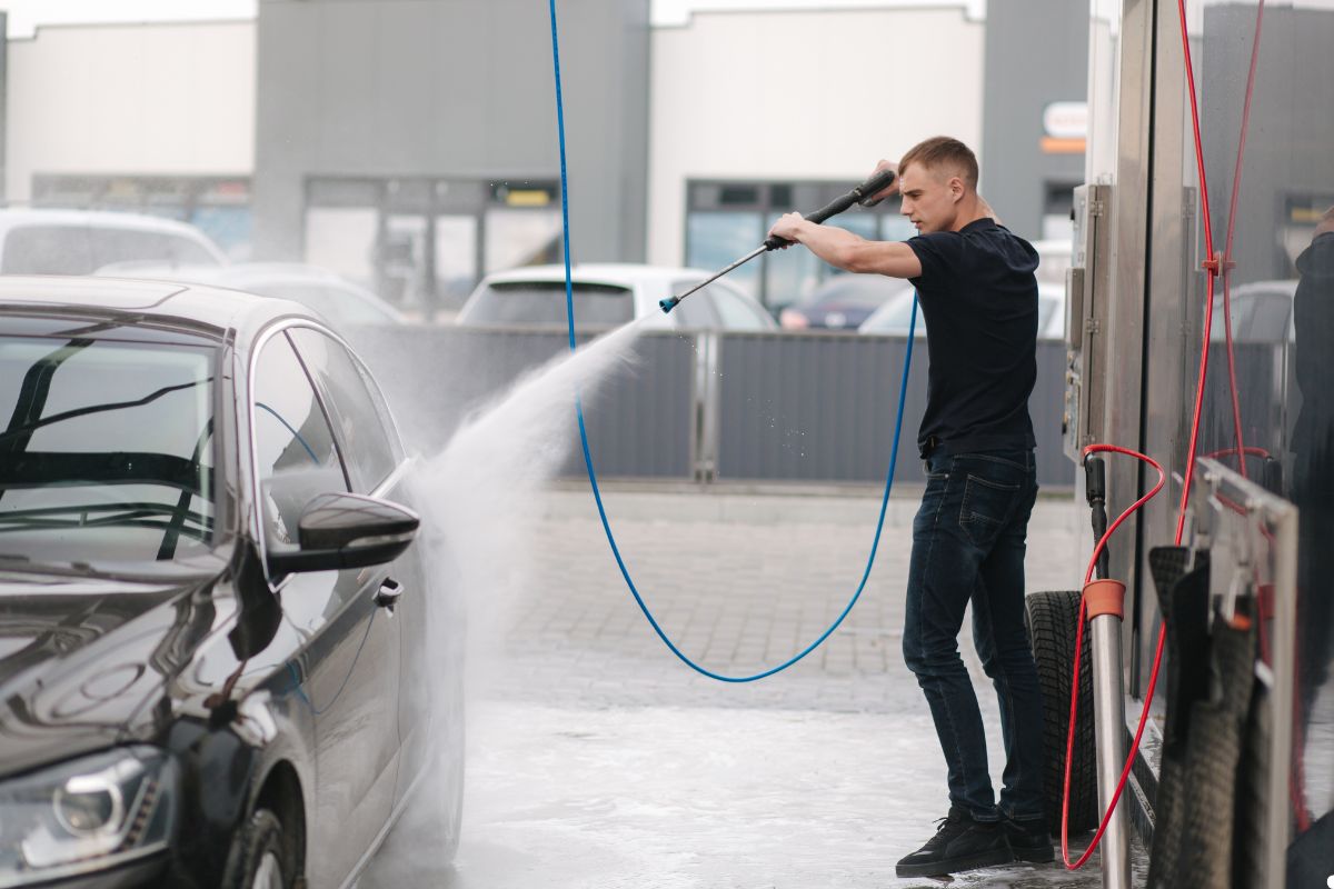 A man washes his car with a hose, spraying water over the vehicle in a sunny outdoor setting.