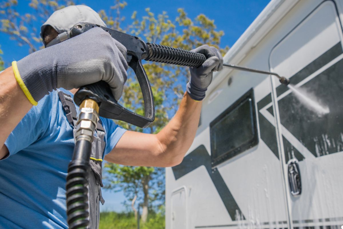 A man uses a pressure washer to remove dirt from the side of an RV, showcasing a bright and clear day.